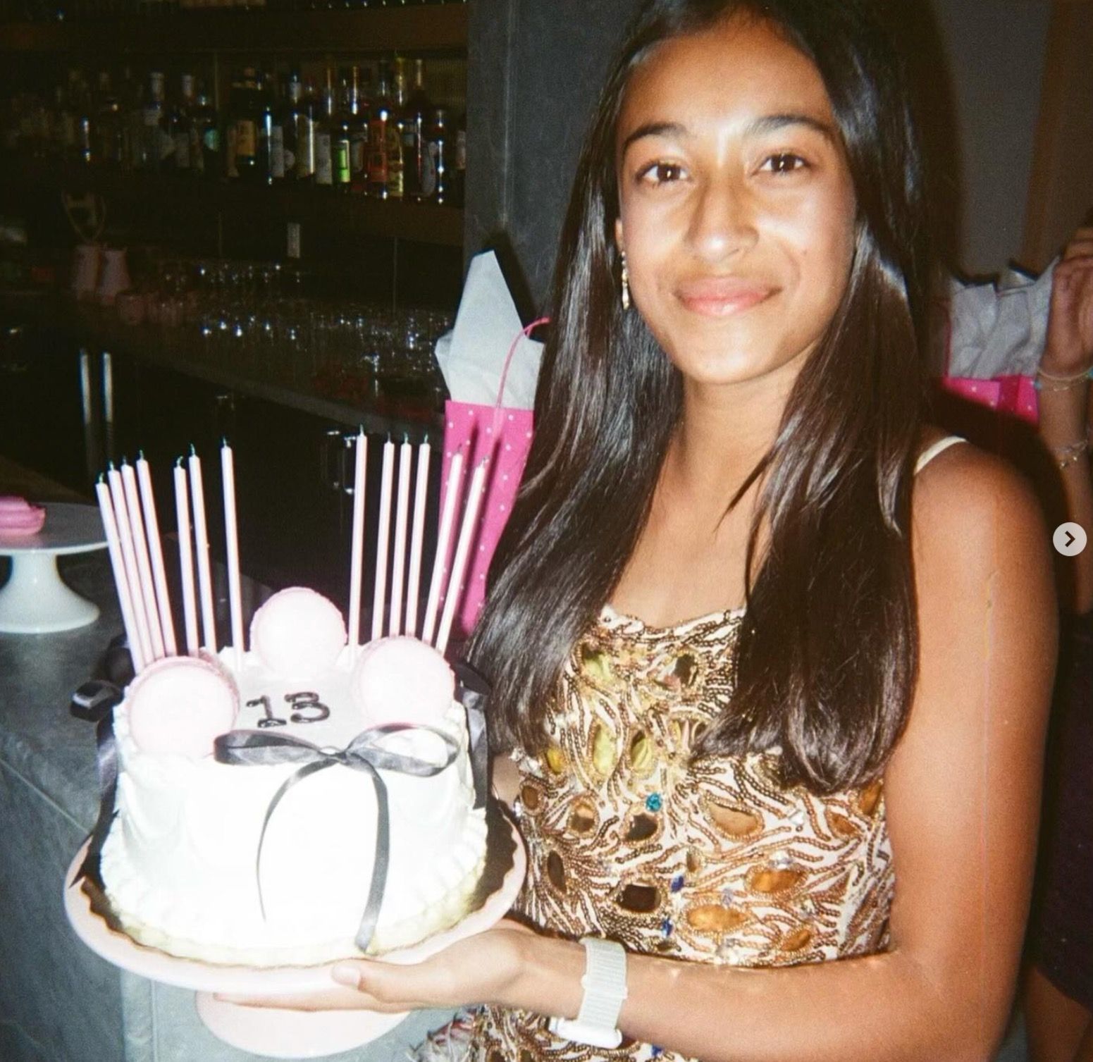 Smiling teen holding a white birthday cake decorated with pink macarons, a black ribbon and tall pink candles with “13” on top, indoor birthday celebration with bar shelves in the background.