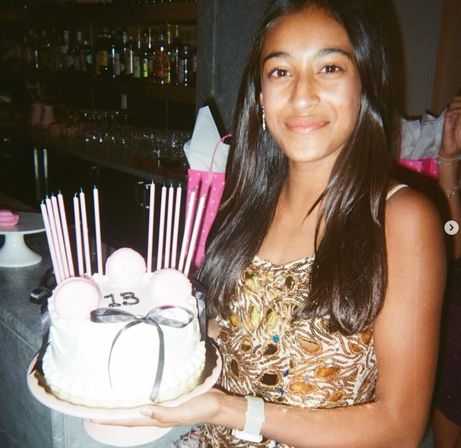 Smiling teen holding a white birthday cake decorated with pink macarons, a black ribbon and tall pink candles with “13” on top, indoor birthday celebration with bar shelves in the background.