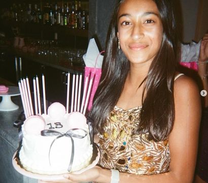 Smiling teen holding a white birthday cake decorated with pink macarons, a black ribbon and tall pink candles with “13” on top, indoor birthday celebration with bar shelves in the background.