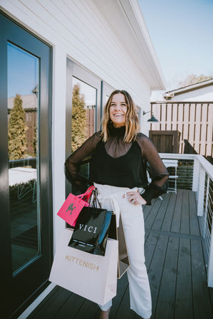 Smiling woman on a modern backyard deck holding colorful shopping bags, wearing a sheer black blouse and white high-waist pants — bright, stylish outdoor shopping moment.