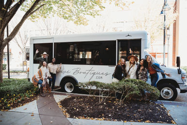 Group of smiling women in fall outfits posing by a white branded boutique shuttle on a downtown street with autumn trees, ready for a shopping outing.