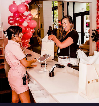 Smiling customer receives a white shopping bag from a cheerful sales associate at a boutique checkout counter with pink balloon decorations