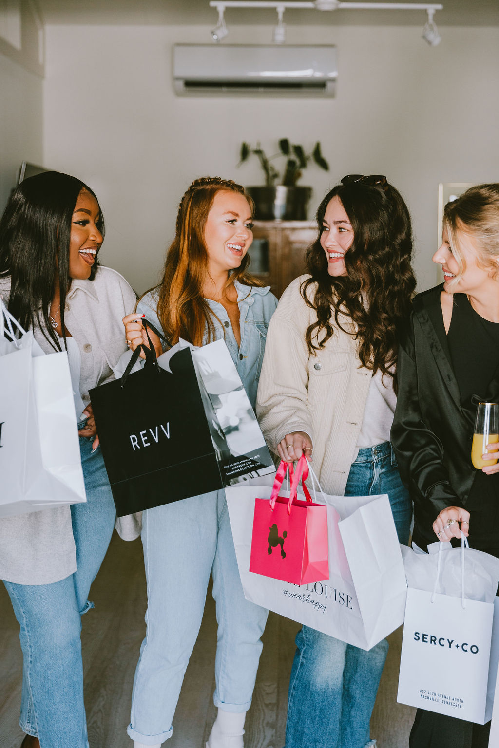 Four smiling women friends laughing indoors while holding colorful shopping bags and a drink after a boutique shopping spree, dressed casually in jeans and light jackets.