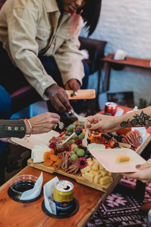 Hands reaching for a colorful charcuterie board — cubes of cheese, brie, grapes, dried apricots, cured meats and jam on wooden serving boards at a cozy indoor gathering with drinks.