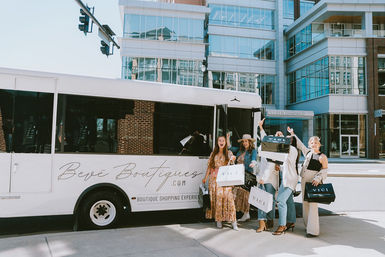 Four stylish women holding shopping bags boarding a white boutique shuttle on a sunny downtown street with modern glass buildings — cheerful boutique shopping outing.