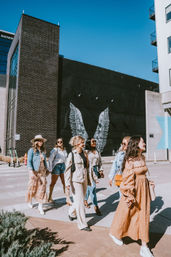 Group of fashionable women walking across a sunny downtown crosswalk in front of a black building with a white angel wings mural, wearing dresses, denim jackets, sunglasses and casual spring outfits