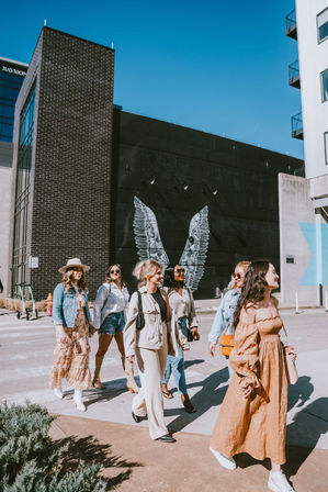 Group of fashionable women walking across a sunny downtown crosswalk in front of a black building with a white angel wings mural, wearing dresses, denim jackets, sunglasses and casual spring outfits