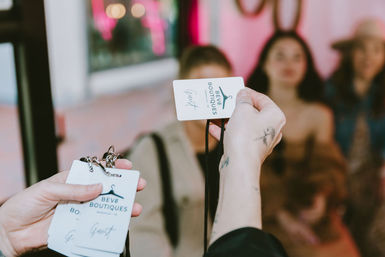 Close-up of hands exchanging white guest-pass lanyards in a bright boutique, with three blurred shoppers in the colorful background.