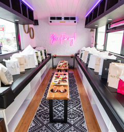 Pink-lit party bus interior with neon 'You Fancy!' sign, black leather banquettes, central wooden table with grazing boards and gift bags for a chic mobile event.