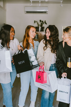 Four smiling women friends laughing indoors while holding colorful shopping bags and a drink after a boutique shopping spree, dressed casually in jeans and light jackets.