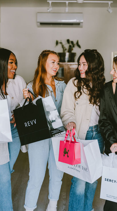 Four smiling women friends laughing indoors while holding colorful shopping bags and a drink after a boutique shopping spree, dressed casually in jeans and light jackets.