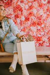 Fashionable shopper in a light denim outfit seated on a bench against a pink rose wall, holding a white shopping bag stuffed with leopard-print tissue, wearing white ankle boots and a tan crossbody bag.