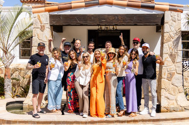 Group of adults raising cocktails on a sunny patio in front of a Mediterranean-style villa with stone columns, terracotta roof tiles, and a palm tree, celebrating a cheerful vacation