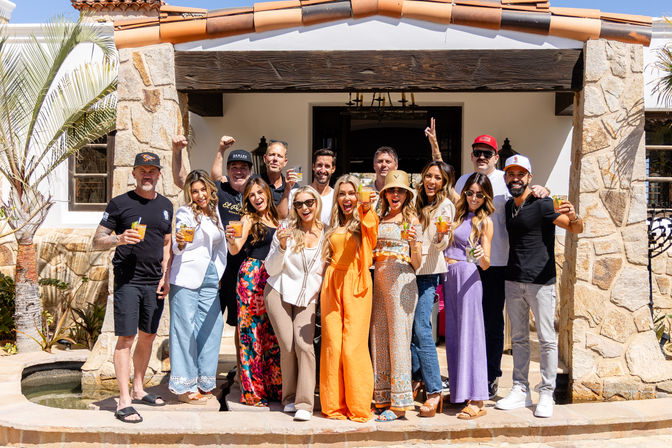 Group of adults raising cocktails on a sunny patio in front of a Mediterranean-style villa with stone columns, terracotta roof tiles, and a palm tree, celebrating a cheerful vacation