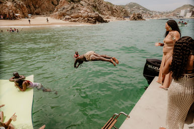 Person mid-dive from a motorboat into green coastal water while friends watch from a dock and floating mat, sandy beach and rocky cliffs in the background—sunny boat party vibe.