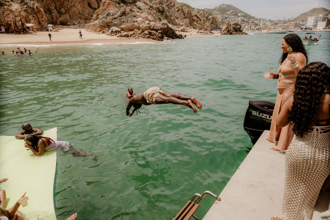 Person mid-dive from a motorboat into green coastal water while friends watch from a dock and floating mat, sandy beach and rocky cliffs in the background—sunny boat party vibe.