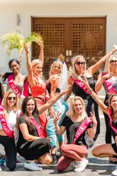 Bachelorette party at a sunny tropical resort — bride in a veil and "Final Fiesta" headband with friends wearing pink sashes, posing cheerfully by a wooden entrance and palm tree.