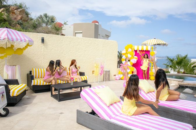 Poolside bachelorette party on a coastal terrace — pink-and-yellow striped loungers and umbrellas, balloon arch, women in matching pink and yellow outfits celebrating with drinks and ocean view