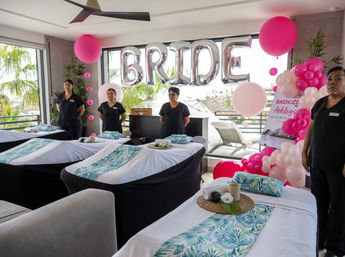 Coastal bachelorette spa setup with four massage tables dressed in tropical-print runners, attendants in black uniforms, large silver 'BRIDE' balloons and a pink balloon arch by floor-to-ceiling windows with palm trees visible outside.