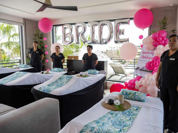 Coastal bachelorette spa setup with four massage tables dressed in tropical-print runners, attendants in black uniforms, large silver 'BRIDE' balloons and a pink balloon arch by floor-to-ceiling windows with palm trees visible outside.