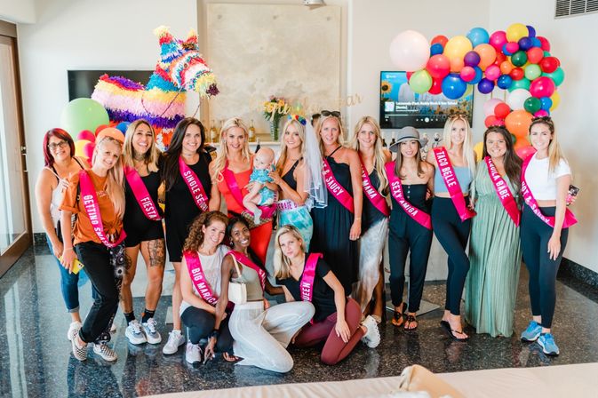 Indoor bachelorette party group wearing pink sashes and a bride veil, posing by a colorful balloon arch, rainbow donkey piñata and drinks