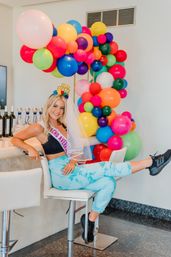 Smiling woman wearing a veil and 'Final Fiesta' headband on a barstool in a modern indoor bar area, tie-dye joggers and black sneakers, surrounded by a vibrant rainbow balloon arch and wine bottles for a bachelorette-style party