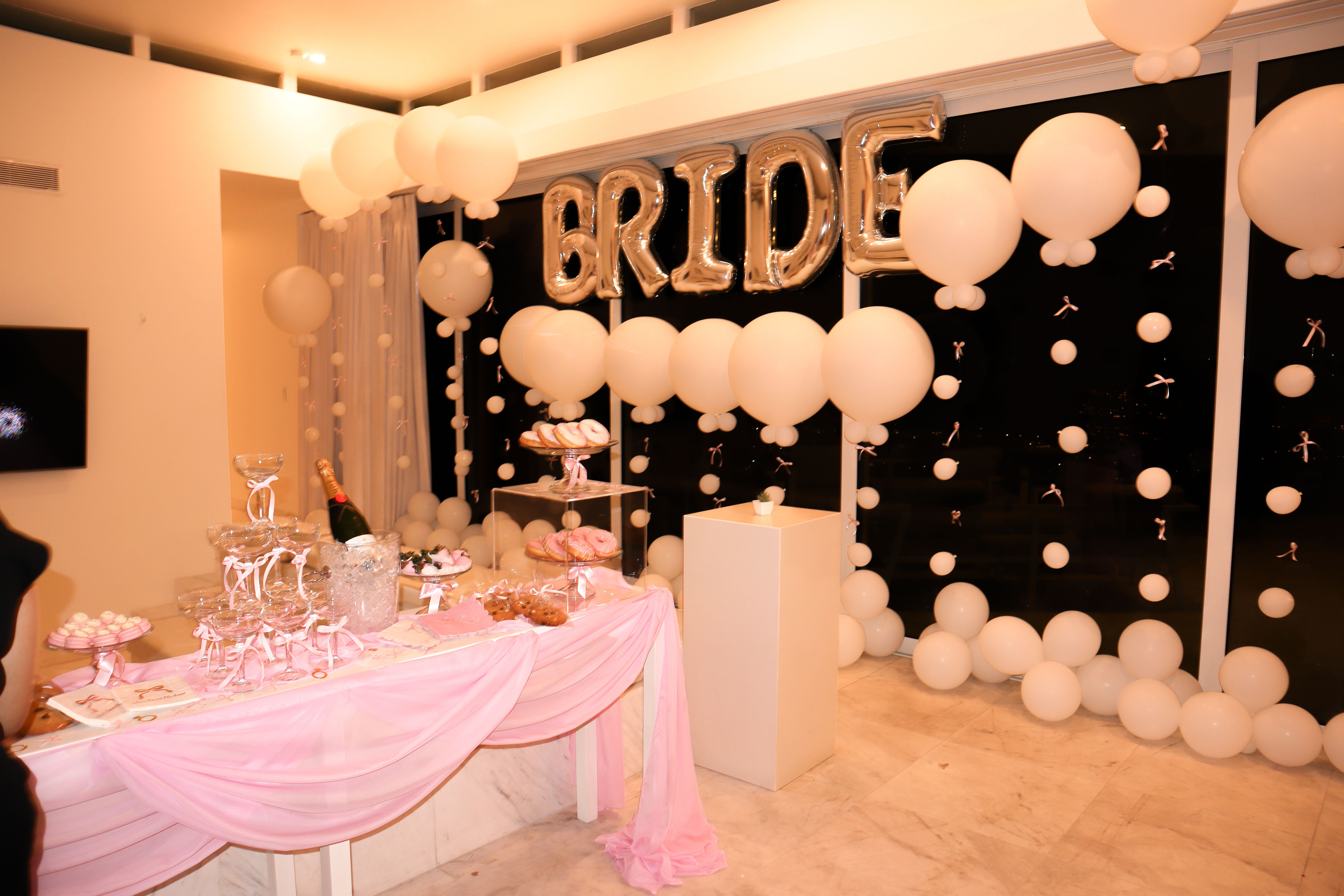 Bridal shower dessert table indoors with silver "BRIDE" balloon letters, white balloon garlands, pink draped tablecloth, champagne bucket and trays of donuts and sweets