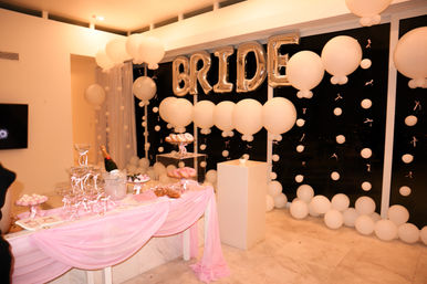 Bridal shower dessert table indoors with silver "BRIDE" balloon letters, white balloon garlands, pink draped tablecloth, champagne bucket and trays of donuts and sweets