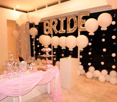 Bridal shower dessert table indoors with silver "BRIDE" balloon letters, white balloon garlands, pink draped tablecloth, champagne bucket and trays of donuts and sweets