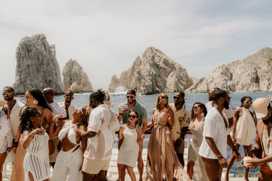 Diverse group dancing and enjoying drinks on a sunlit boat with the iconic rock arch of Cabo San Lucas, Mexico rising from the blue sea