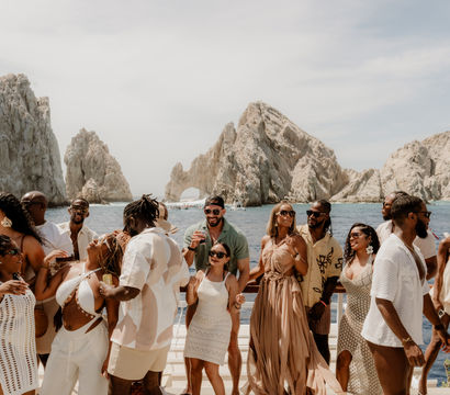 Diverse group dancing and enjoying drinks on a sunlit boat with the iconic rock arch of Cabo San Lucas, Mexico rising from the blue sea