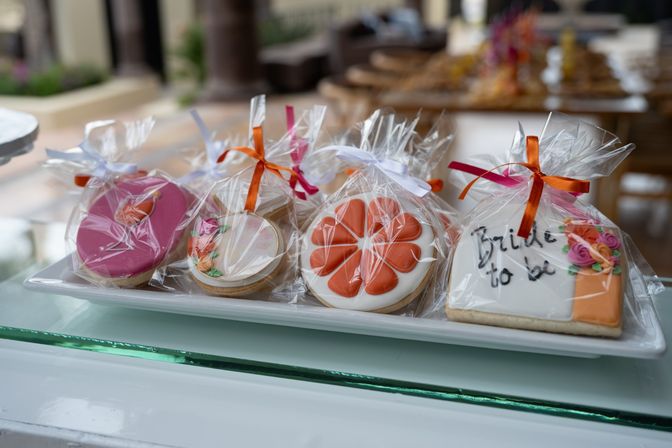 Platter of decorated sugar cookie party favors wrapped in clear cellophane with orange and pink ribbons — floral, heart and an iced “Bride to be” cookie, perfect bridal shower treat