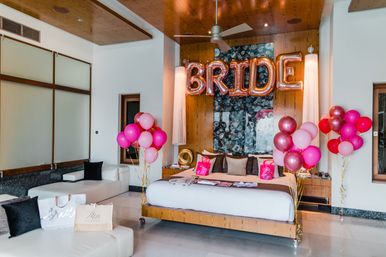 Modern hotel bridal suite decorated for a bachelorette with rose gold BRIDE balloons above a king bed, pink and red balloon clusters, gift bags and white lounge seating
