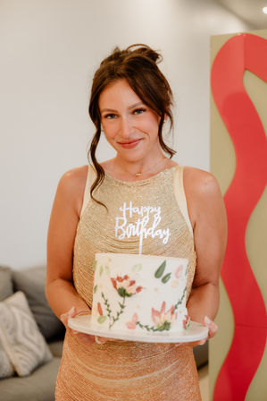 Smiling woman in a shimmery gold dress holding a round cake decorated with painted flowers and a "Happy Birthday" topper in a cozy indoor living room setting