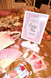 Playful pink bachelorette party spread on an indoor wooden table: framed 'Entry Fee 1 Shot' sign, heart-shaped pink sunglasses, pink fan, face-cutout stickers, mini bottle, spa eye masks and assorted bridal shower favors.