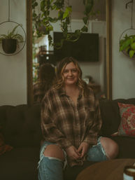 Smiling woman sitting cross-legged on a tufted sofa in a cozy living room, wearing a plaid shirt and ripped jeans, surrounded by hanging houseplants, a mirror and colorful throw pillows