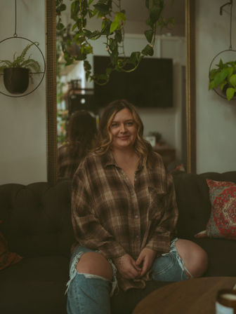 Smiling woman sitting cross-legged on a tufted sofa in a cozy living room, wearing a plaid shirt and ripped jeans, surrounded by hanging houseplants, a mirror and colorful throw pillows