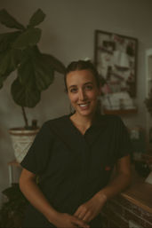 Smiling woman with a nose ring and tied-back hair leaning on a wooden counter in a cozy, plant-filled home studio with warm moody lighting and a pinboard in the background.