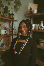 Portrait of a smiling person with dreadlocks and colorful beaded necklaces in a cozy boho pottery shop surrounded by plants and wooden shelves