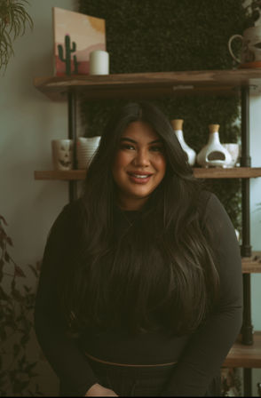 Smiling woman with long dark hair and nose ring in a black top, posed indoors against wooden shelves with ceramics, plants and a small cactus painting, cozy portrait vibe.