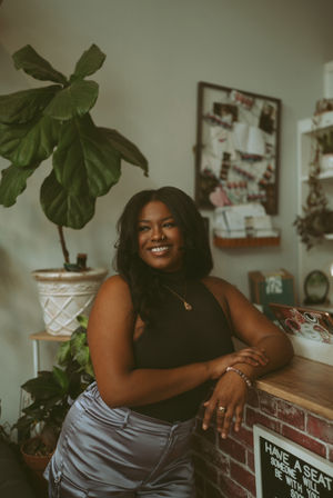 Portrait of a smiling woman leaning on a brick counter in a cozy, plant-filled indoor studio, wearing a black sleeveless top and satin pants in warm natural light.