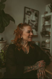 Smiling red-haired woman with floral neck tattoo and intricate hand tattoos, wearing a black top and multiple rings, seated at a wooden counter in a cozy, plant-filled indoor studio with shelves blurred in the background.