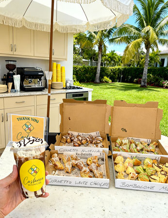 Iced chocolate coffee with whipped cream held over an outdoor patio counter of open boxes of Italian pastries (cornetti, bomboloni), an espresso station, fringe umbrella and palm trees in a tropical backyard.