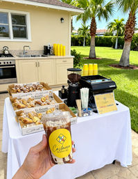 Hand holding iced coffee in front of an outdoor coffee bar with espresso machine, grinder, stacks of yellow cups and trays of croissants on a white tablecloth, palm-tree backyard