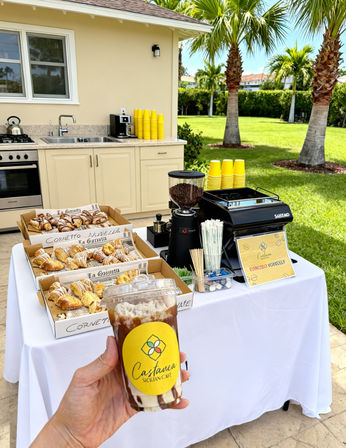 Hand holding iced coffee in front of an outdoor coffee bar with espresso machine, grinder, stacks of yellow cups and trays of croissants on a white tablecloth, palm-tree backyard