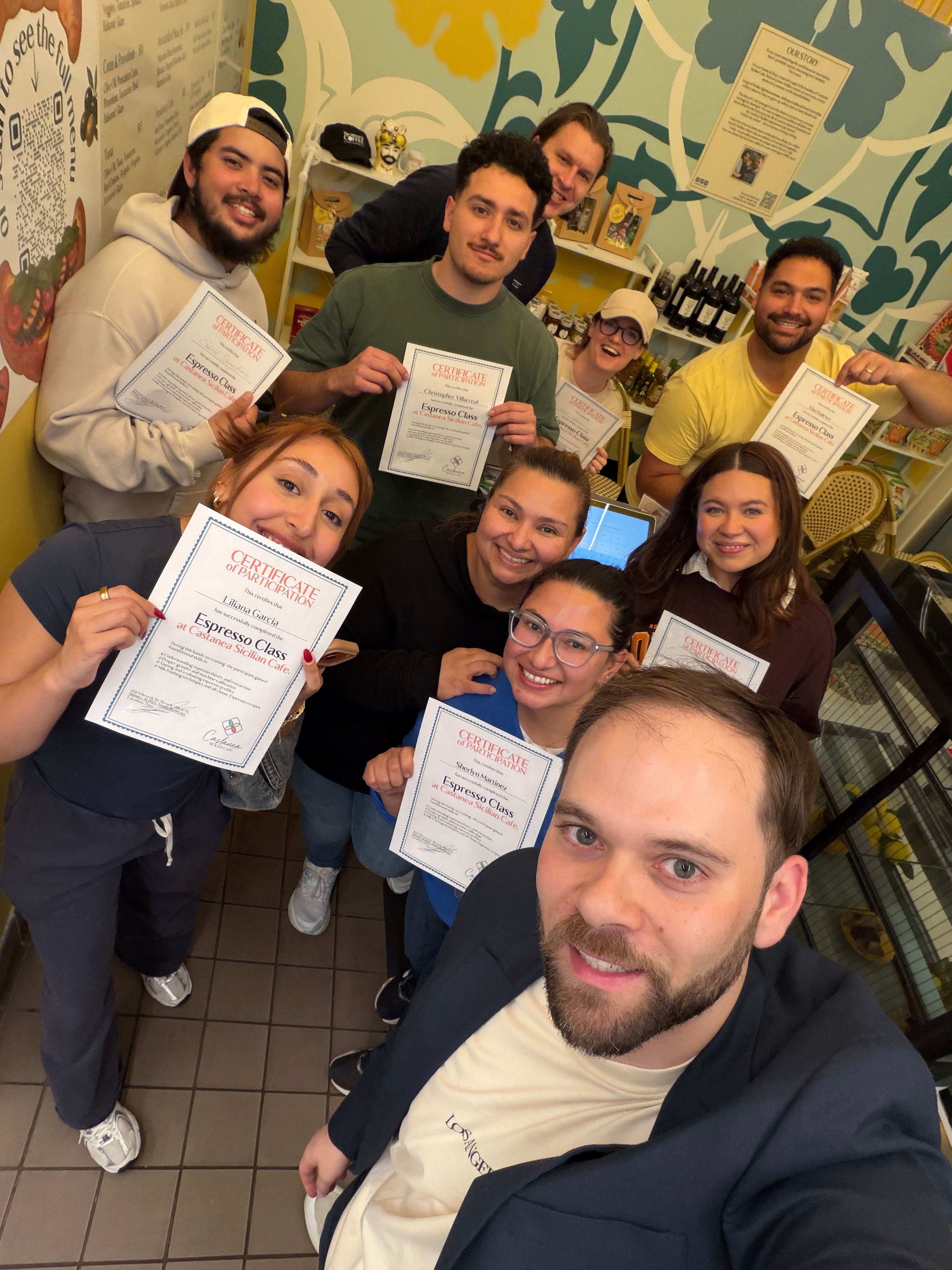 Smiling group selfie of nine people in a bright, patterned coffee shop holding certificates after an espresso class.