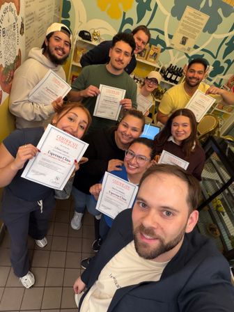 Smiling group selfie of nine people in a bright, patterned coffee shop holding certificates after an espresso class.