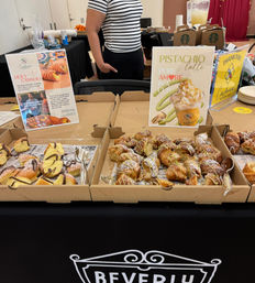 Bakery sampling table with trays of cannoli and pistachio‑drizzled pastries and a 'Pistachio latte' sign at an indoor food tasting event