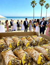 Close-up box of ricotta cannoli with yellow custard, powdered sugar and chocolate chips in foreground, oceanfront beach wedding cocktail reception with guests in formal attire, palm trees and waves under a bright blue sky