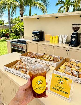Iced coffee in a yellow-labeled to-go cup held in front of an outdoor coffee bar with espresso machines, stacks of yellow cups, trays of Italian pastries (cornetti and bomboloni), and palm trees on a sunny tropical patio.